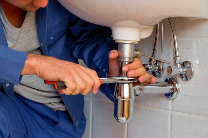 Plombier professionnel installant des tuyaux sous un lavabo, avec des outils et des raccords, dans une salle de bain moderne.